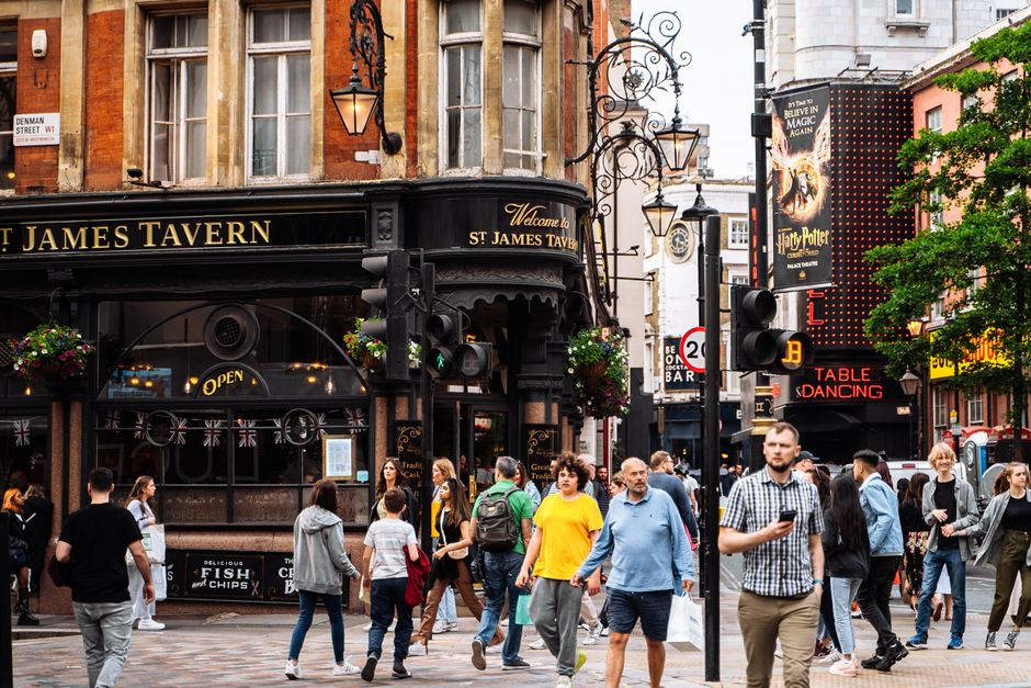 A busy urban street scene in front of the historic St James Tavern with its black-painted facade and decorative hanging lanterns. The cobblestone pavement is filled with pedestrians, some walking acro